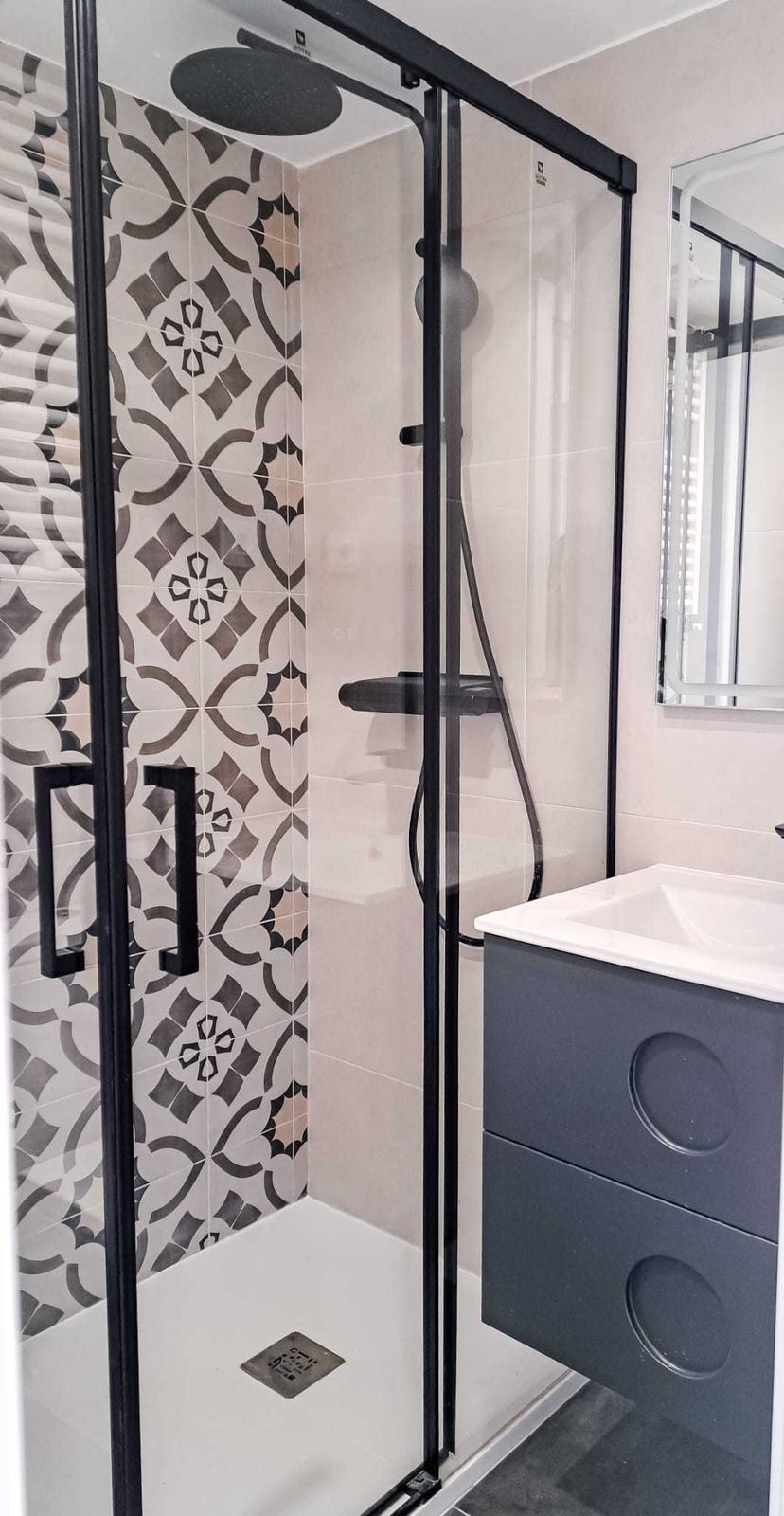 Ducha con azulejo de aire hidráulico en gris y crema, mampara de perfil negro, rociador de techo y mueble de lavabo gris en el baño de una reforma integral de 80 m² en Delicias, Arganzuela